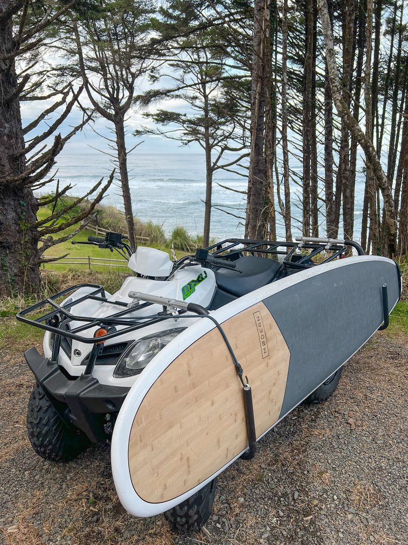 White ATV Quad shown holding a white and blue SUP Stand Up Paddle board.  The vehicle is parked at the top of a hill overlooking the ocean.