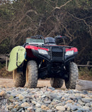 Red Honda all terrain vehicle all wheel drive with a green surfboard attached to the side of the ATV via surf rack.  The motor vehicle is on top of a pile of rocks near some trees.