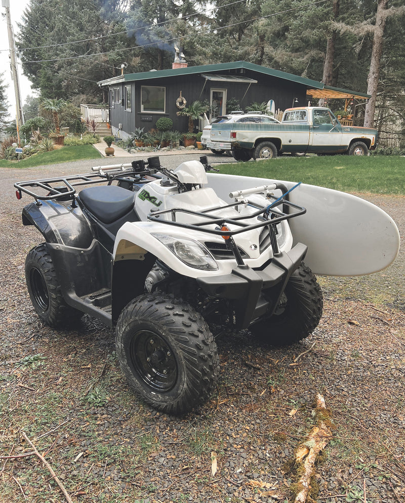 White ATV Quad shown in front of a house.  The ATV is holding a white surfboard using a surf rack mounted to the motorbike. 