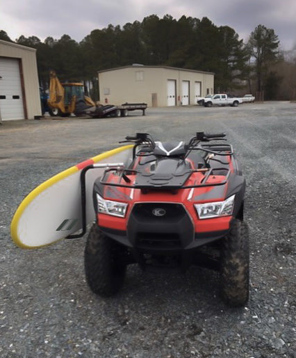 Quad and ATV Surfboard rack shown mounted on a Red All Terrain Vehicle with a yellow longboard attached to the side.  There are buildings in the background along with white trucks, construction equipment, and trees. 