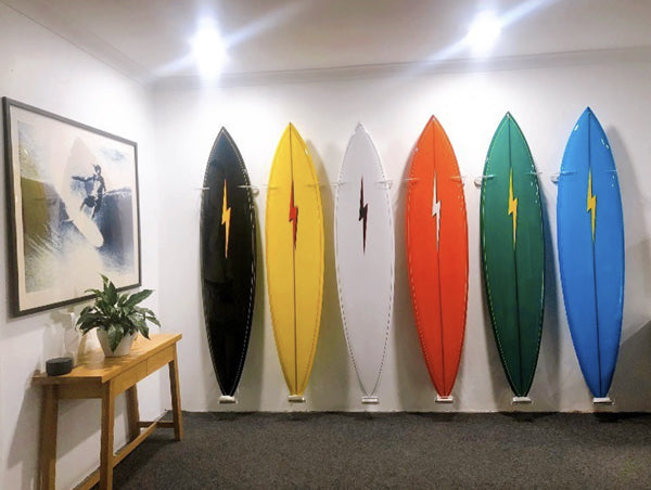 Several surfboards are shown mounted next to one another in a board room using the acrylic surfboard vertical longboard racks.  The surfboards are various colors from blue, green, red, white, yellow, and black.