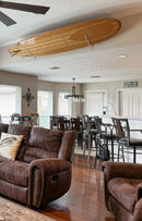 Living room and kitchen shown with a wooden longboard surf board mounted near the ceiling using a clear acrylic wall rack.  Brown leather sofa's in the foreground.