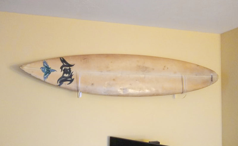 Acrylic trophy style wall rack shown on a yellow wall holding a brown NEV surfboard above a television.
