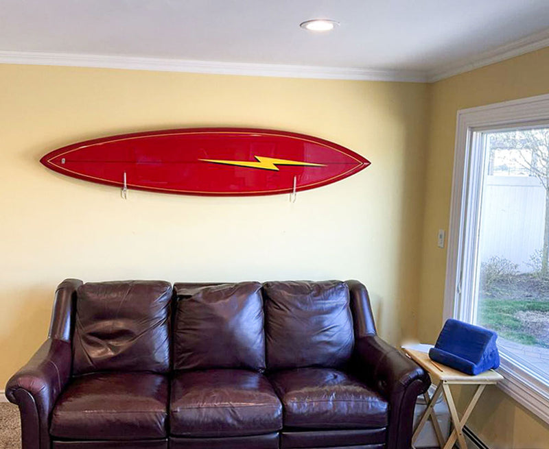 Acrylic Flush rack with a red Jerry Lopez surfboard mounted above a leather couch next to a window.