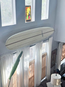 Clear acrylic surfboard wall rack shown holding a new triple stringer longboard above and below high windows, on display in a living room. 