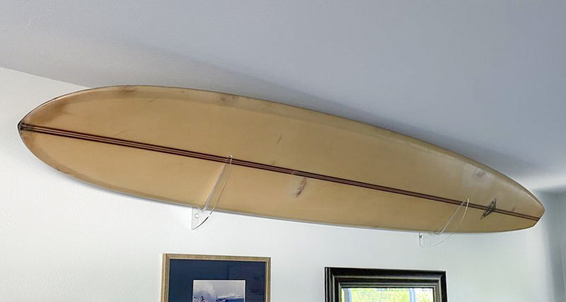 clear acrylic surfboard wall rack holding a retro longboard.  The board is beige & brown in color.  It is hung on a white wall with some picture frames below the board.