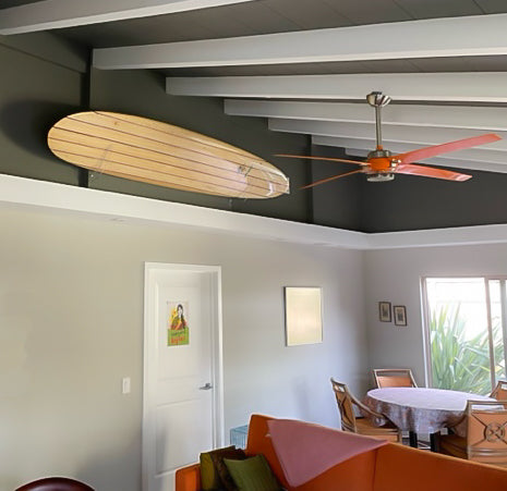 Clear acrylic surfboard wall rack that is holding a longboard.  The rack is holding the board at a downward 45 degree angle.  There is a vaulted ceiling with a dark grey and white beams.  There is a table and couch in the foreground. 