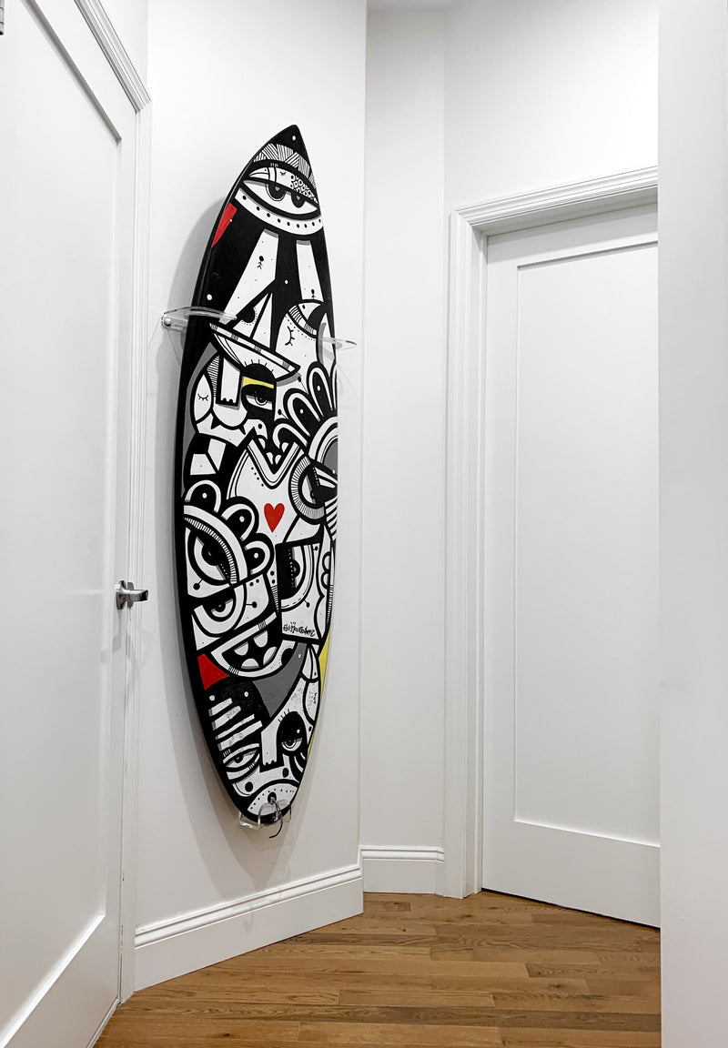 Surfboard with vibrant graphics being displayed in a hallway with wooden floors.  The board has several images on it with eyes in black and white with hints of red and yellow.  The surfboard is mounted to the wall in the vertical position using clear wall hangers.