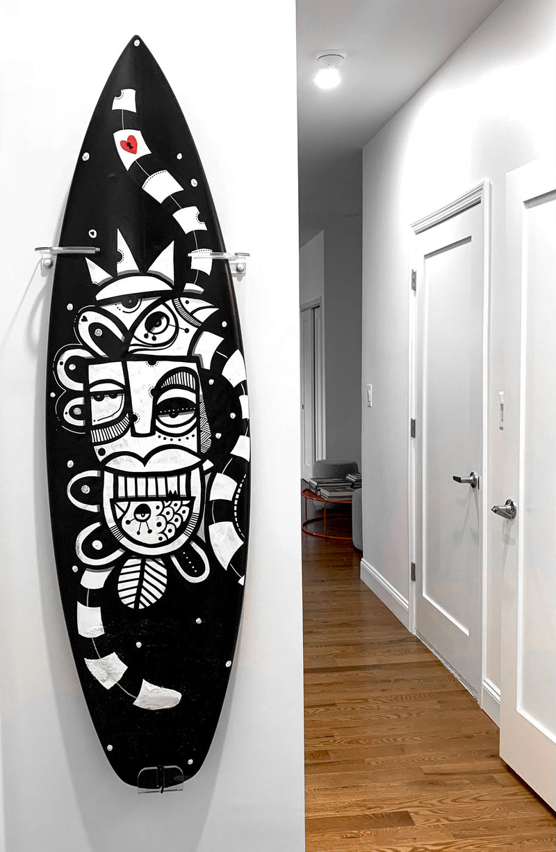 Surfboard mounted vertically to a wall, in the hallway of a home with wooden floors. The surfboard displays black and white art. 