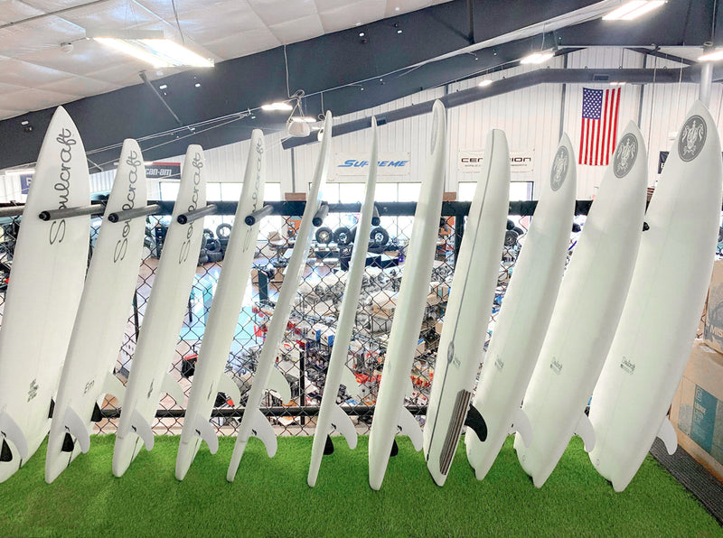 Front view of the vertical surfboard wall rack made of black metal  mounted to a fence overlooking a boating showroom floor.  The surf racks are holding several white wakesurf boards in the vertical position.  The boards are laying on fake grass turf.