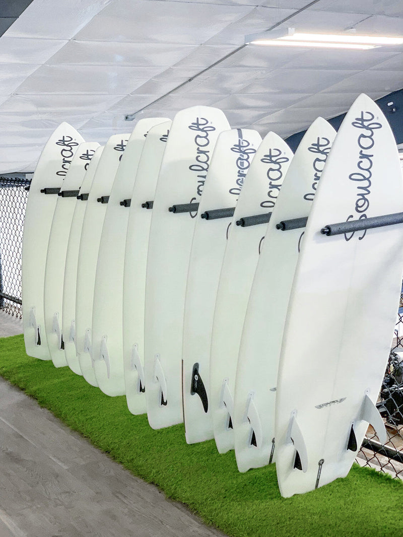 Black metal vertical surfboard wall rack holding several Wake Surf Boards in the vertical upright position.  The boards are laying on top of faux grass.