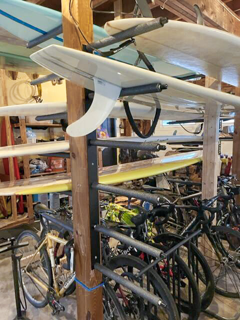 black metal wall rack mounted to some wooden beams in the middle of a garage.  The board racks are holding several longboards, and the racks are mounted directly over several bicycles. 