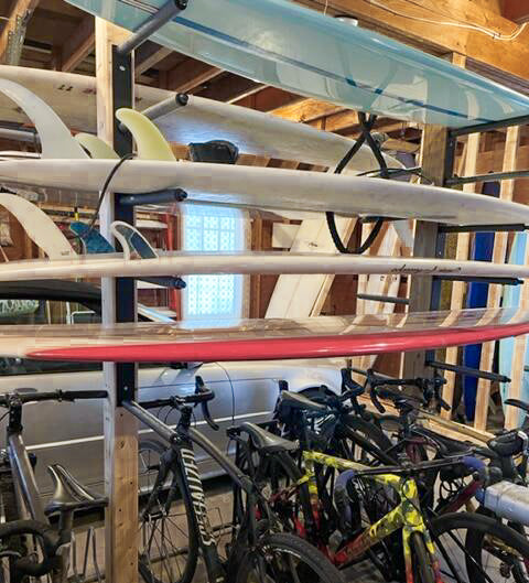 Black metal wall rack mounted to beams in a garage.  A silver car is in the background of the garage.  There are several bikes mounted below the surfboards. 