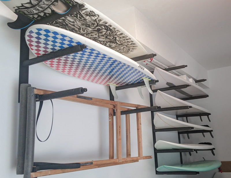Two black metal surfboard wall racks mounted side by side next to each other on a white wall. The racks are holding several high-performance surfboards.