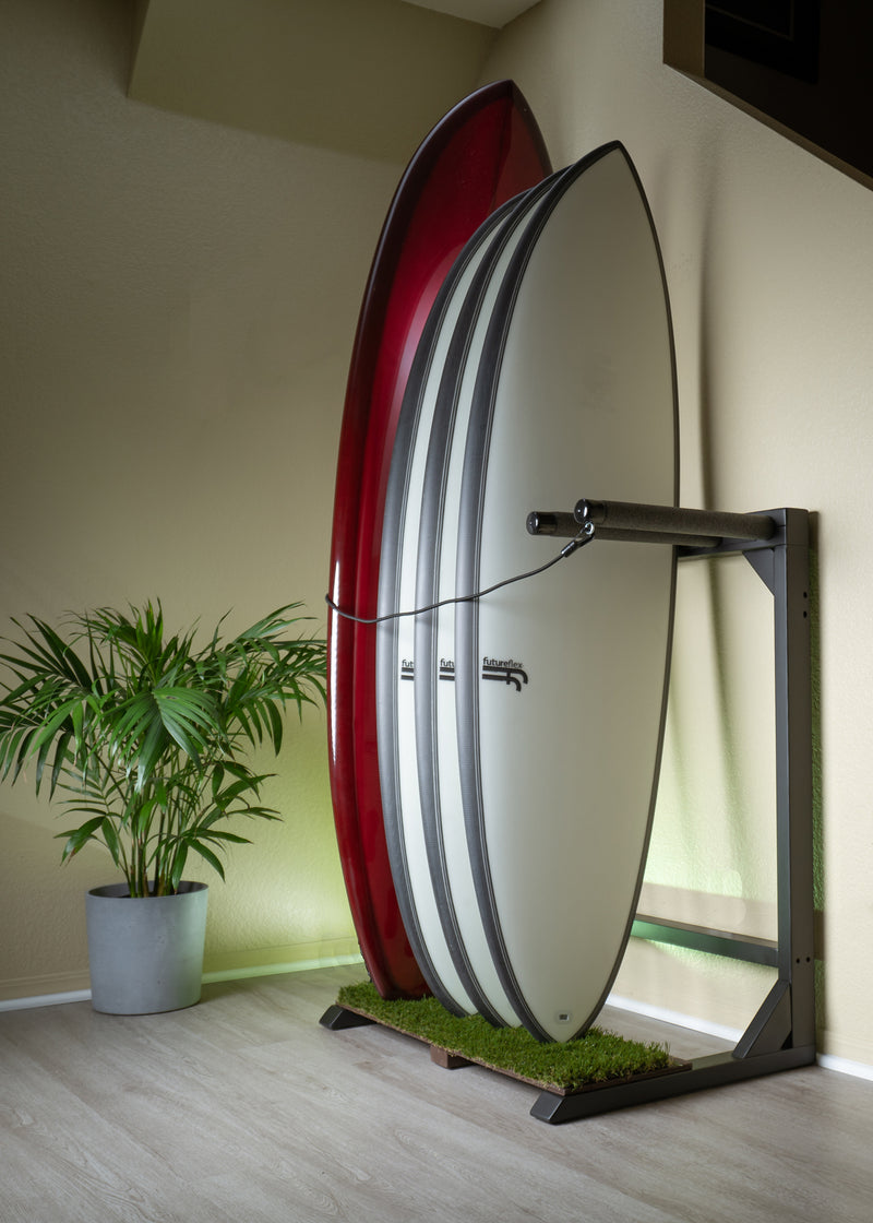 Vertical freestanding surfboard rack shown in a a room next to a green plant with a green flashing light in the background.  The vertical surf rack is flat black in color, has a grass base where the surfboards are.  The rack is holding 4 surfboards.