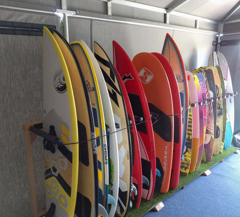 Several Grassy surfboard racks shown next to one another holding numerous wake boards, and surfboards.  There is a blue carpet on the ground.