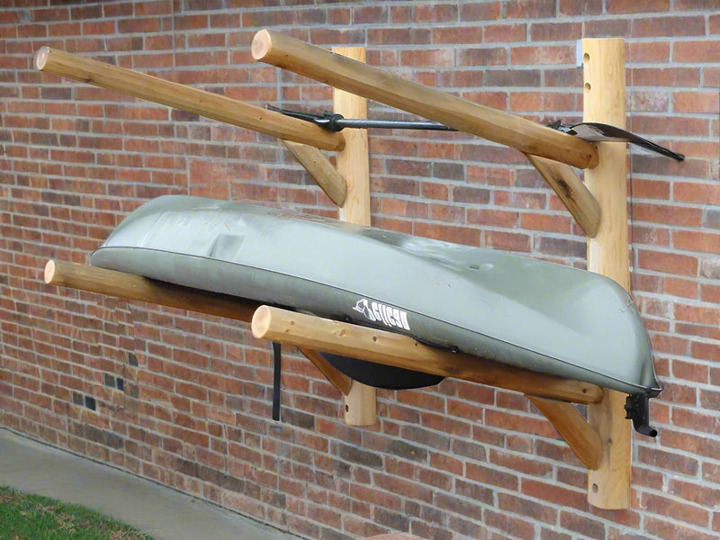 Kayak shown on a wooden log kayak rack mounted to a brick wall with some grass underneath. 