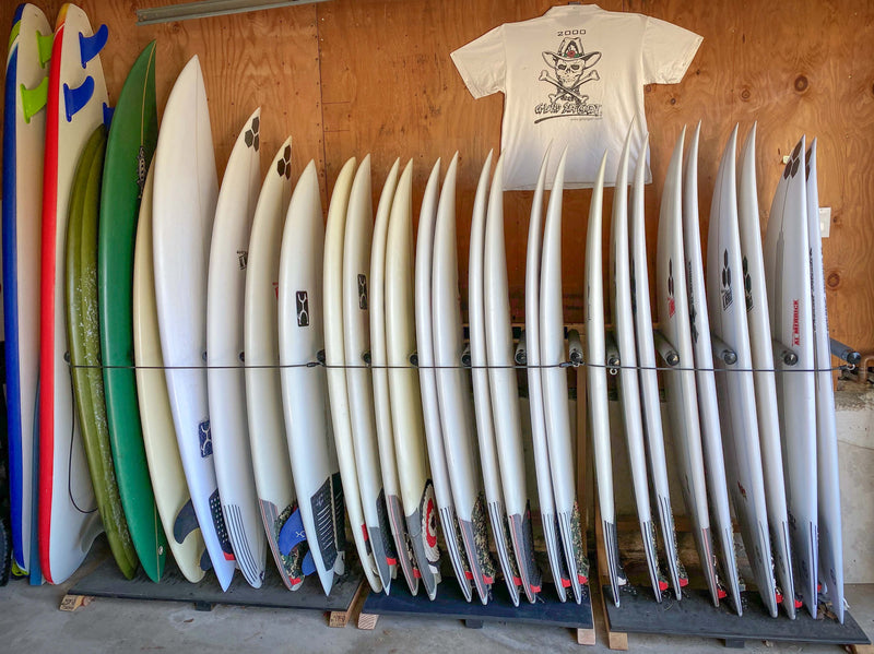 Three custom foamy freestanding rack shown holding many shortboards.  The racks are custom that holds two surfboards per slot.  There is a wood wall in the background with a t-shirt mounted above the surfboards. 
