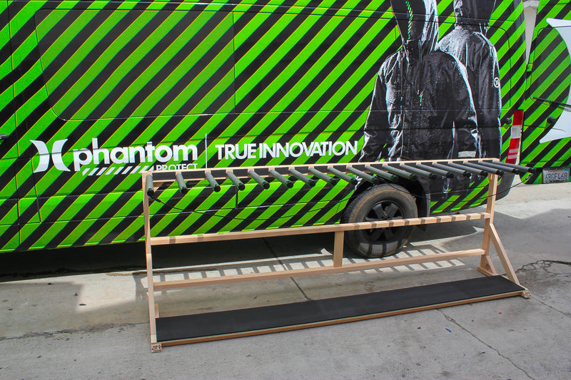 Custom freestanding vertical surfboard rack for the hurley pro.  The empty rack has slots to hold up to 18 surfboards in the vertical position.   In the background there is a van with Hurley branding that says Phantom projects and True Innovation with the picture of a jacket.  The van is painted in green and black stripes. 