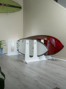 White SUP and Surfboard Dock Rack shown holding 2 surfboards next to a staircase inside a home. Another green surfboard is mounted on the wall in the background with some plants on the floor and in the foreground. There is grey wooden floors.