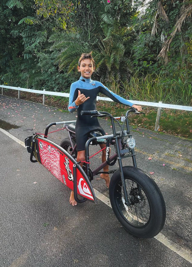 E-bike Surfboard Rack -Dual-C Clamp Model shown with a young man on the North Shore of Oahu, holding a red board.