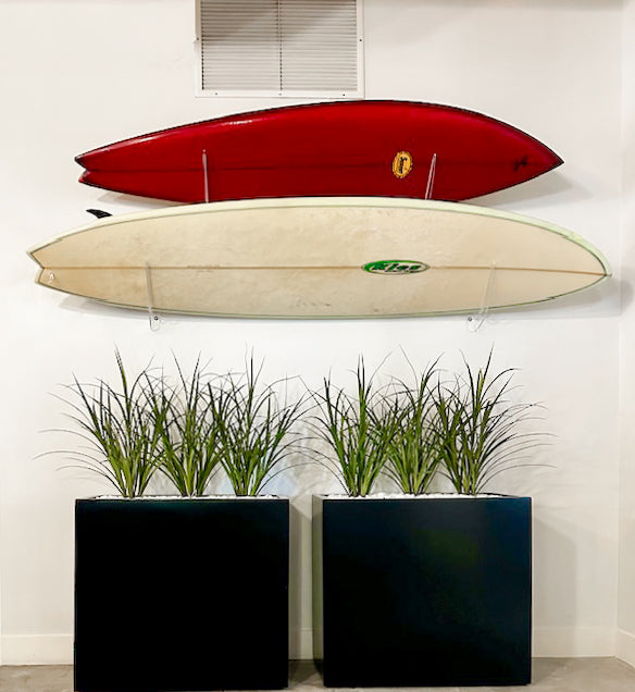 Two acrylic surfboard wall racks holding surfboards above two plants.  The boards are being held in the horizontal position, and are angled off the wall at 45 degrees.The top board is red and the bottom board is white. 