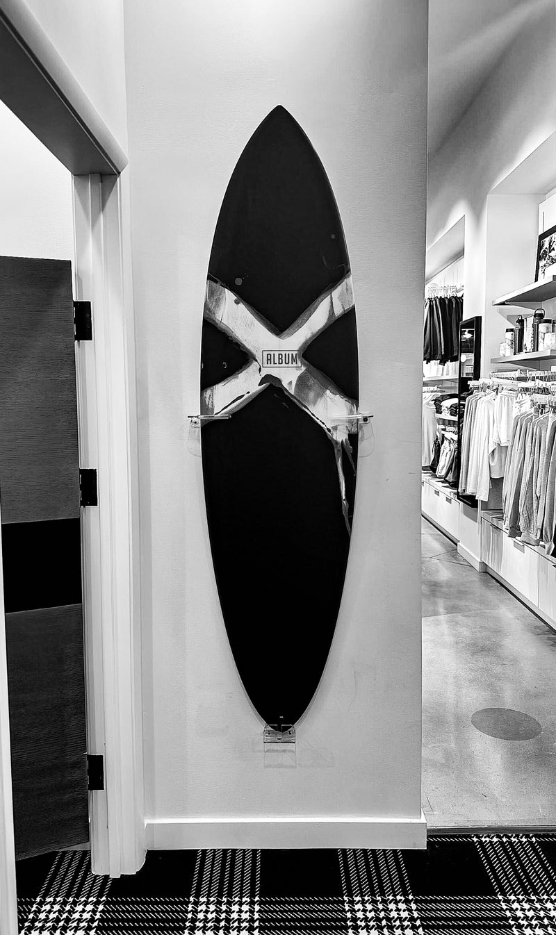 Album surfboard mounted to the wall with acrylic surfboard wall mounts in a retail setting.  There are several clothes racks in the background and a dressing room to the side. 