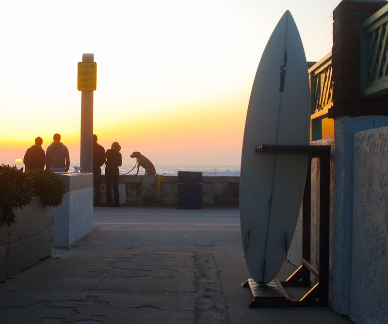 Picture of a sunset with the silhoutte of an epic surf racks vertical freestanding rack in the foreground, laying next to a wall.There is a group of people in the background next to a seawall watching the sunset. 