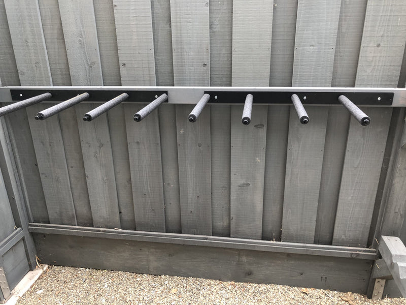 Vertical surfboard wall racks in black metal mounted up against a grey wooden fence.  There are small pebbles on the ground.  The surf racks have grey foam covering and end caps. 