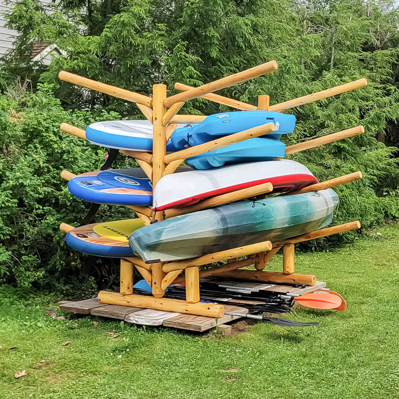 8 Boat Freestanding Log Kayak rack holding multiple Stand Up Paddleboards and several Kayaks and other watercraft with paddles on the small deck below. All sitting on grass with lush green bushes in the background.