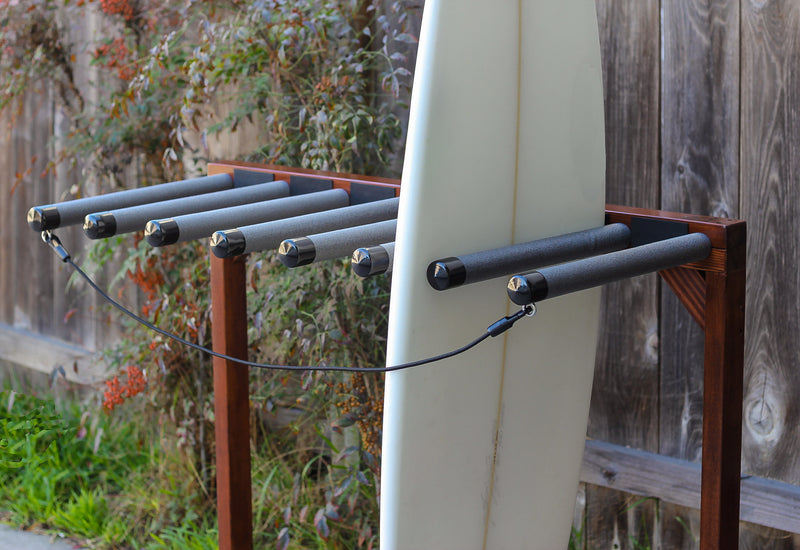 Front angle of the finished foamy surf rack.  The rack is dark brown in color with a high-gloss finish.  There is a white surfboard being held in the rack.  There is a grey fence in the background with several bushes at the bottom of the fence. 