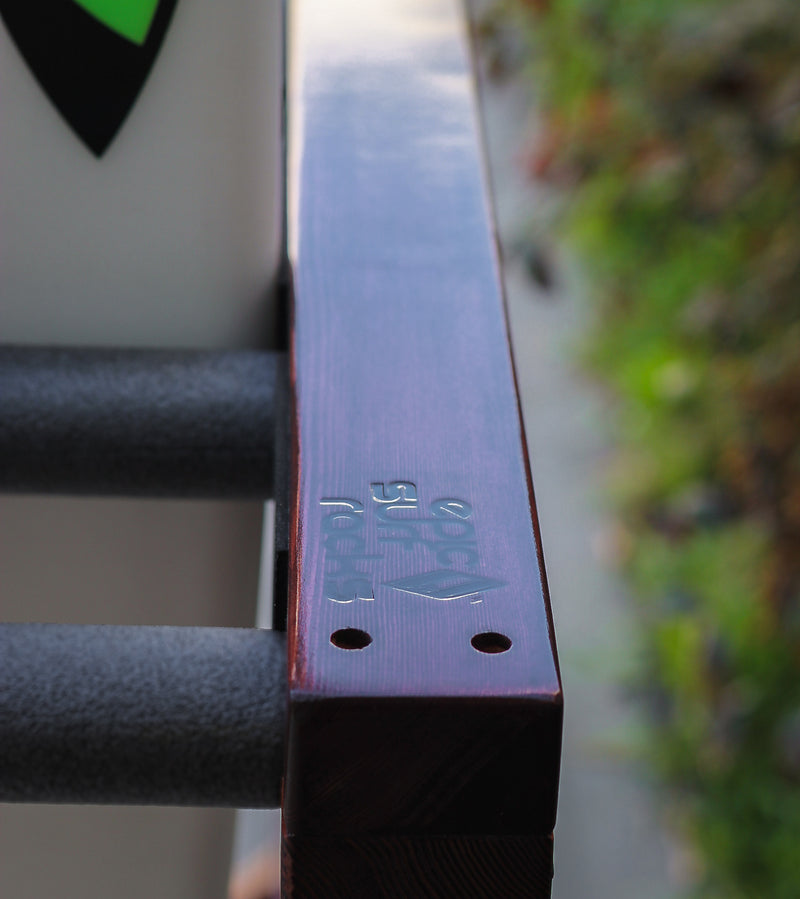Close up detail shot of the top of the finished Foamy surf rack showing the dark wood color and high-gloss finish.  The rack is outdoors with green bushes to the side of the rack. 