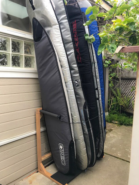 The Foamy freestanding rack shown holding several longboards in a customers backyard.  Each board is in a surfboard travel bag for protection.  There are plants in the background and the surfboard rack is leaning up against a house. 
