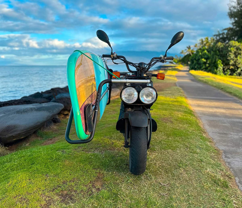 Honda Ruckus holding a Stand Up Paddleboard, near the beach in a tropical environment. There are lots of green trees in the background and a walkway.