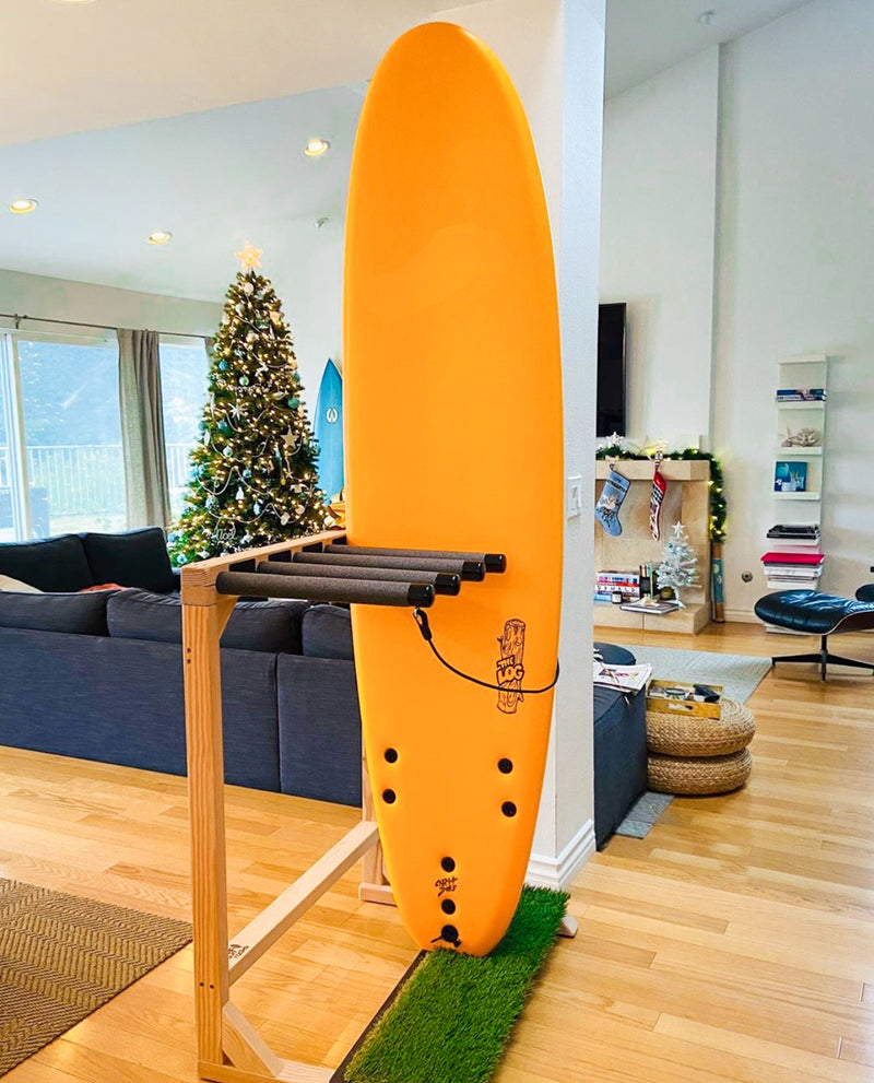 Customer photo of a grassy surf rack holding a brand new orange soft-top surfboard.  There is a couch and christmas tree in the background.