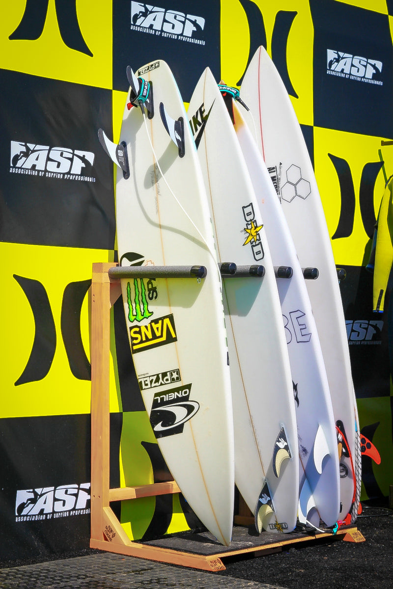 Foamy vertical freestanding surf rack shown holding several pro surfers boards during the Hurley Pro ASP professional surf contest.  The board has 4 white surfboards that are covered in sponsosred stickers.  There is a checkered hurley logo in the back in yellow, with the ASP (association of surfing professionals) mixed in the checkered pattern in black.