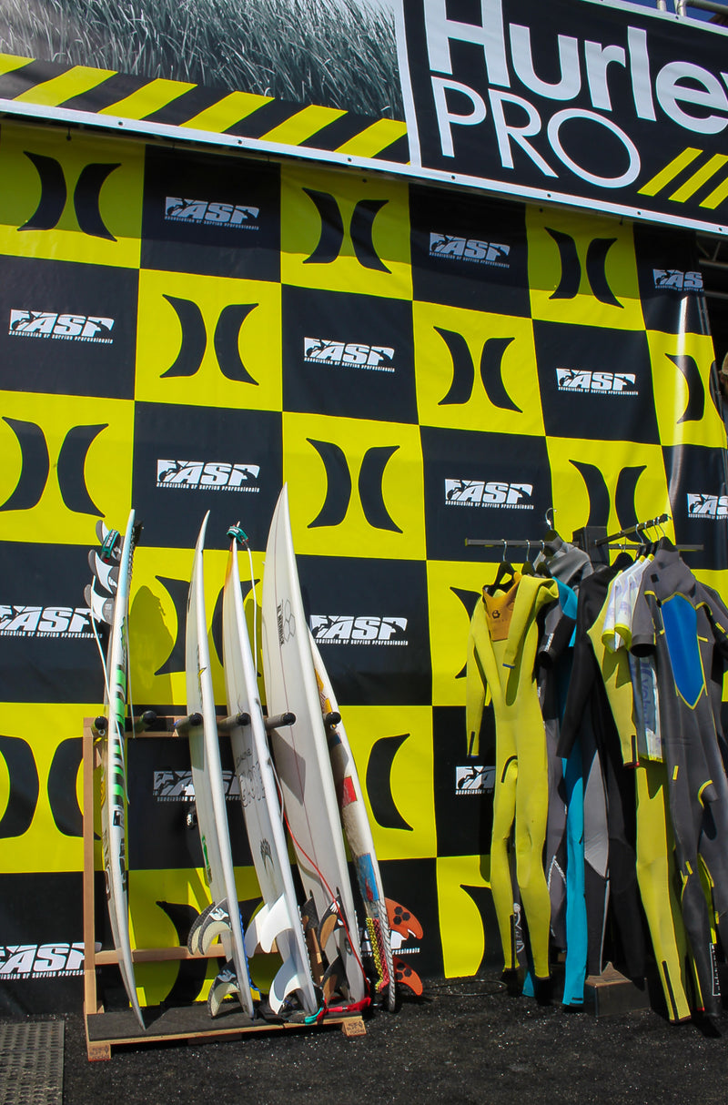 Pulled back shot of the Foamy freestanding surfboard stand holding several professional surfers boards during the Hurley Pro contest. There are several wetsuits on a rack to the right of the board rack.  The hurley pro branding is all around in the background in black and yellow. 