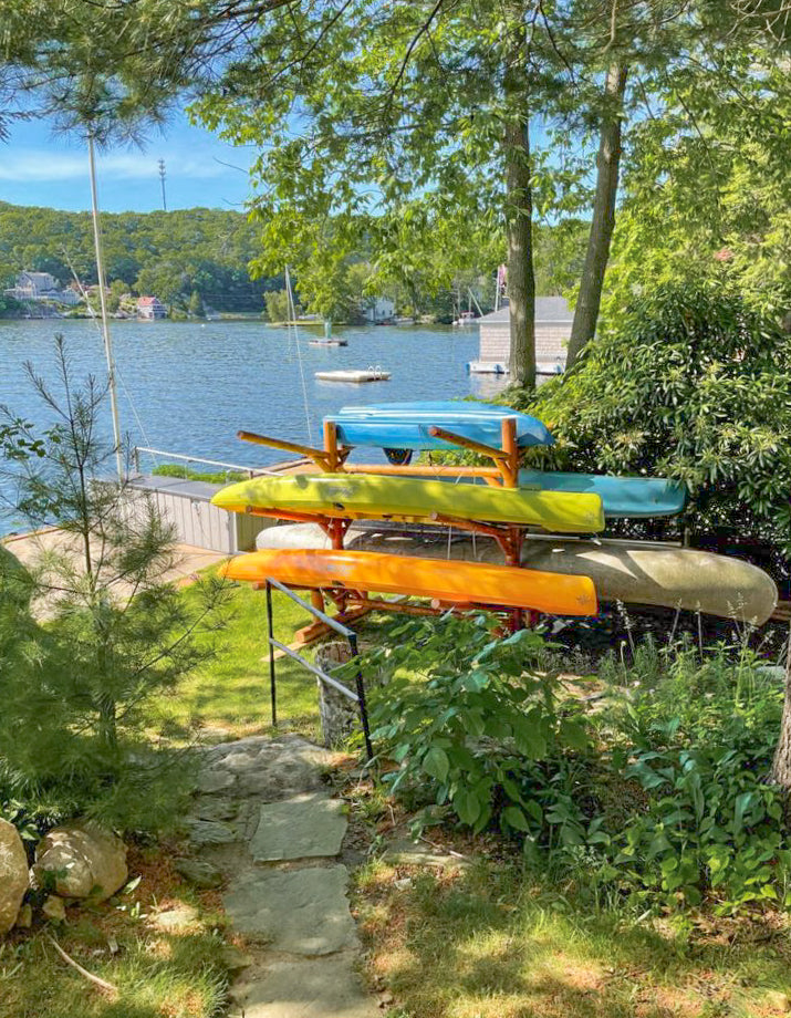 Canyon Brown Log Kayak rack for 6 watercraft holding several canoes and Kayak next to a beautiful lake.  There are lush green trees and bushes in the foreground and background. 