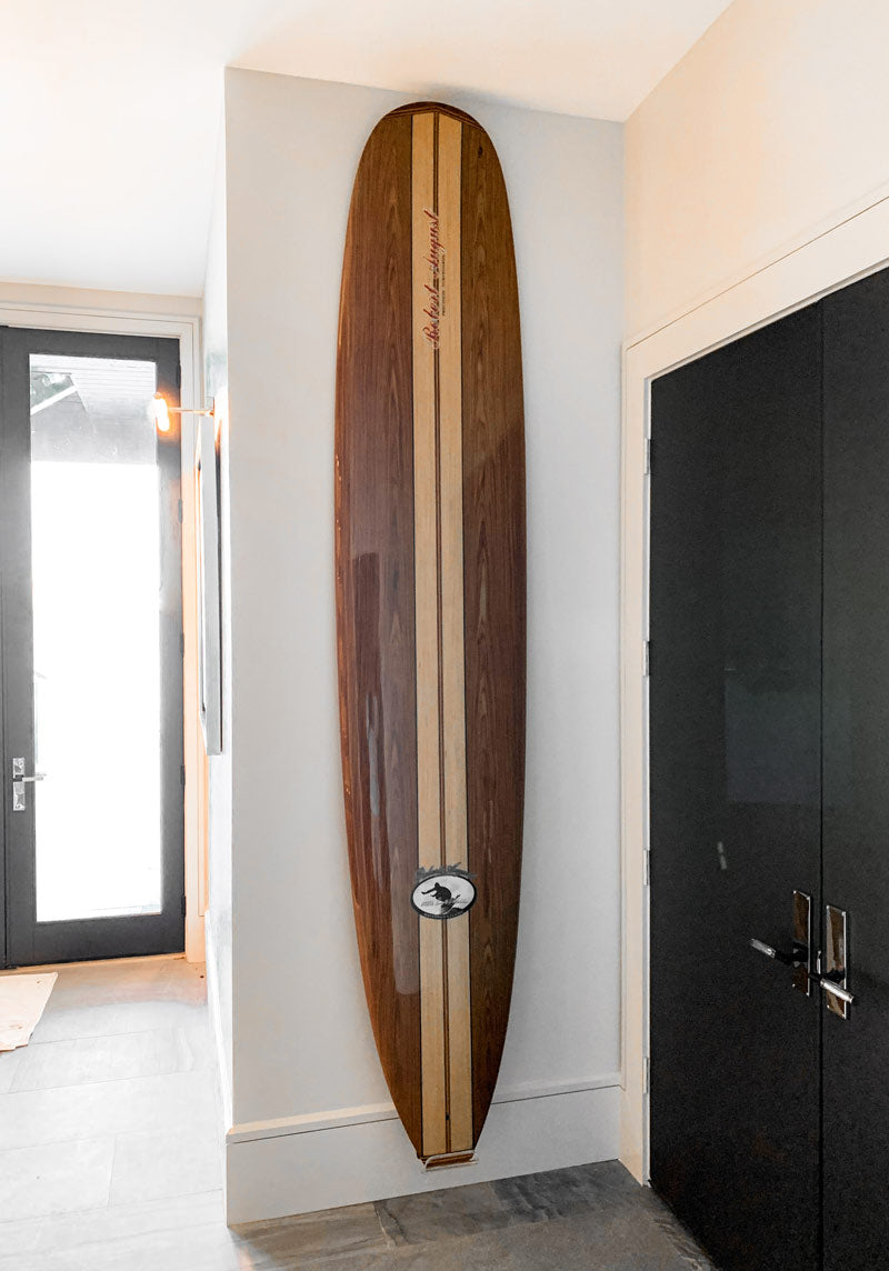 Wooden longboard surfboard mounted to the wall in a hallway using a clear acrylic vertical wall rack. 