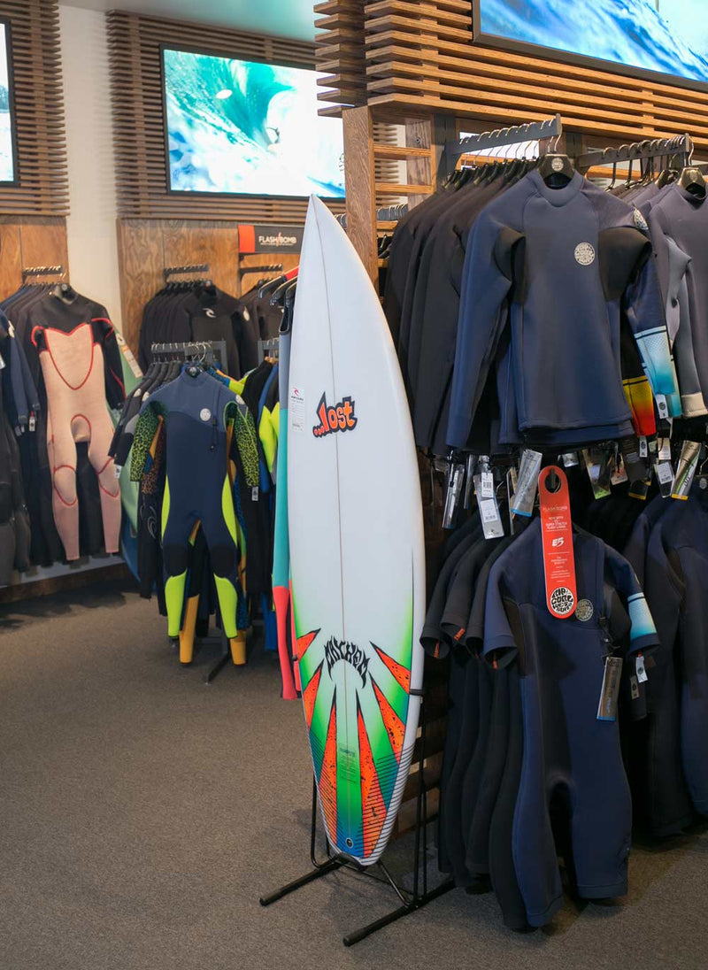 A lost surfboard being shown on display in a surf shop in front of some wetsuits. The board is standing up with the steel single freestanding surf rack.