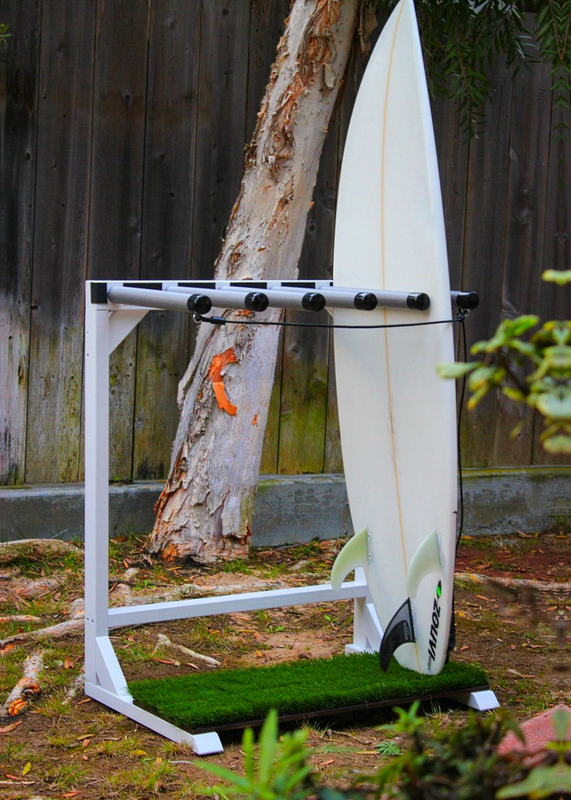 Marshmallow Grassy surf rack shown in white on the side of a yard. This vertical freestanding surfboard rack is holding just a single shortboard. The faux grass base is shown protecting the bottom of the board. There are bushes in the foreground and a tree & fence in the background.