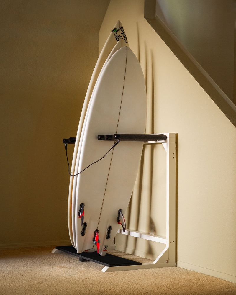 White vertical freestanding rack shown holding multiple surfboards next to a staircase. The rack is in a living room, and has beige walls and carpet.