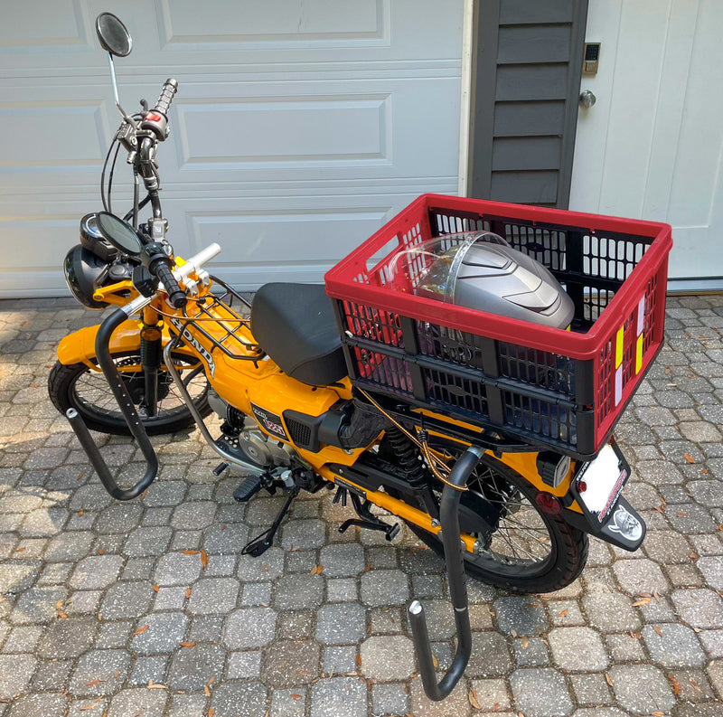 Yellow Honda Moped with a surfboard rack attached, parked in front of a white garage door. The moped has a red basket which is holding a full faced helmet.