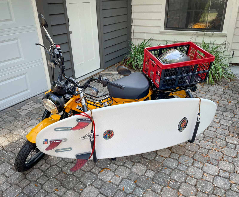 Yellow Honda Moped with a surfboard rack holding a performance surfboard in a backyard. The moped has a bucket in the back holding a helmet.