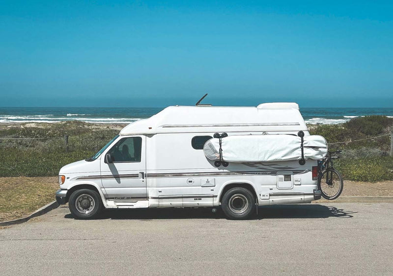 Seasucker rack shown holding a longboard surfboard mounted directly to the side of an RV recreational vehicle parked on the beach. 