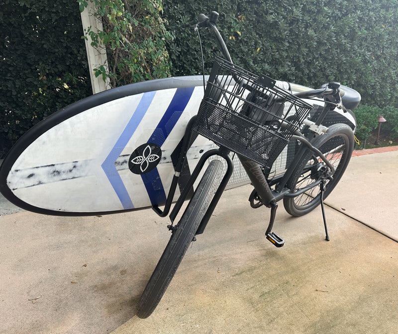 Black beach cruiser bicycle shown parked in a drive way with a white SUP / Stand Up Paddle Board attached to the side of the bike. 