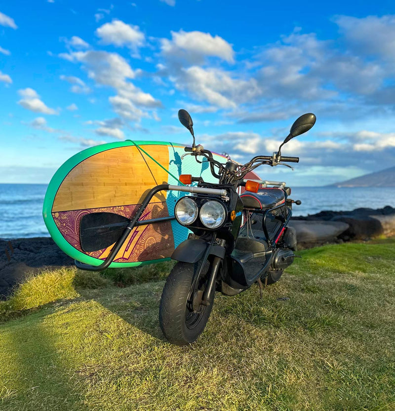 Honda Ruckus with our dual mount SUP moped rack on the grass next to the ocean with a mountain in the background.  The sun is about to set and the sky is exceptionally blue. 