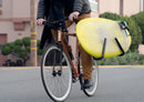 Cyclist on a fixed gear bike riding to the beach with a yellow surfboard attached to his bike with a surf rack.  He has a wetsuit strapped over his shoulder.