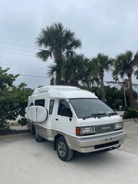 A White Townace camper van is parked with palm trees in the background.  There is a surfboard mounted to the side of the vehicle using suction cup mounts. 