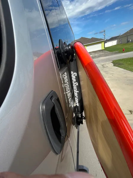 Close up of the suction mount rack mounting a red surfboard to the side of a silver van.  There are houses and a blue sky in the background. 